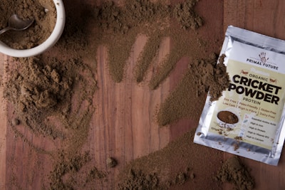 A wooden surface is covered with a light brown powder, with a handprint visible in the scattered substance. A small white bowl filled with the same powder is accompanied by a metal spoon. Nearby, a packet labeled 'Organic Cricket Powder' displays various nutrition labels and claims about the product, such as being soy-free and protein-rich.