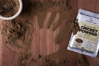 A wooden surface is covered with a light brown powder, with a handprint visible in the scattered substance. A small white bowl filled with the same powder is accompanied by a metal spoon. Nearby, a packet labeled 'Organic Cricket Powder' displays various nutrition labels and claims about the product, such as being soy-free and protein-rich.