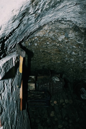 In a dimly lit, rustic cellar, an axe with a yellow and brown handle is propped against a textured stone wall. Below, there are crates filled with various items, possibly wood and other stored goods. The atmosphere is rugged and has an old, well-used feeling.