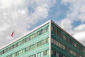 A modern building with a teal-green glass facade, positioned at an angle against a partially cloudy sky. The building has numerous windows and a Canadian flag is mounted at the top.