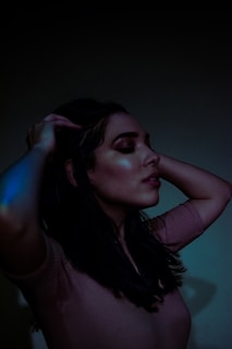 A woman with closed eyes holds her hands in her hair, under dim lighting, creating a serene and contemplative atmosphere. The subtle glow on her skin reflects soft lighting adding to the mood.
