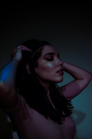 A woman with closed eyes holds her hands in her hair, under dim lighting, creating a serene and contemplative atmosphere. The subtle glow on her skin reflects soft lighting adding to the mood.