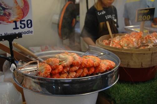 A food stand displays large cooked shrimp in a metal bowl and cooked crabs in a wooden container. There are price signs indicating the cost per gram for each type of seafood. A woman can be seen behind the stand assisting with sales.