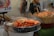 A food stand displays large cooked shrimp in a metal bowl and cooked crabs in a wooden container. There are price signs indicating the cost per gram for each type of seafood. A woman can be seen behind the stand assisting with sales.