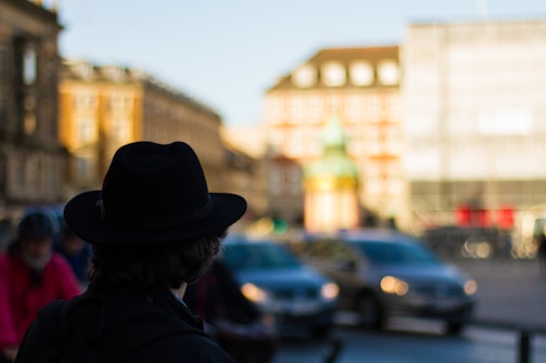 A person wearing a black hat is seen from behind in a city setting. The background is blurred, showing a mix of architectural buildings and moving cars, with a bright, sunny atmosphere.