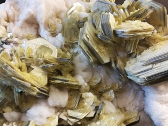 A collection of pale yellow, hexagonal crystalline formations clustered together against a backdrop of white, cotton-like mineral deposits. The crystals have a shiny, reflective surface and are layered in a staggered pattern, creating a complex, textured appearance.