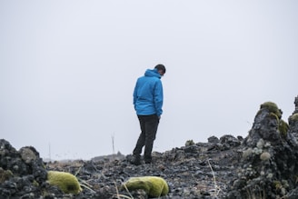 A model wearing a moss green technical jacket standing on a rocky Mourne mountain peak under overcast skies.