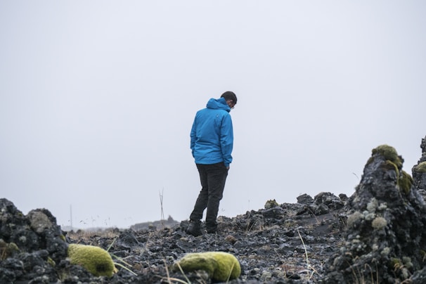 A model wearing a moss green technical jacket standing on a rocky Mourne mountain peak under overcast skies.