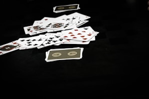 A scattered pile of playing cards lies on a dark surface, displaying various suits including hearts, clubs, spades, and diamonds. The scene conveys a sense of casual disarray or the aftermath of a card game.