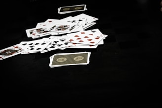A scattered pile of playing cards lies on a dark surface, displaying various suits including hearts, clubs, spades, and diamonds. The scene conveys a sense of casual disarray or the aftermath of a card game.