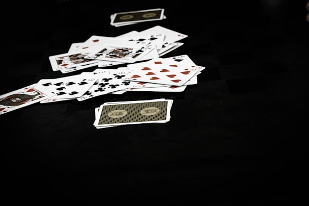 A scattered pile of playing cards lies on a dark surface, displaying various suits including hearts, clubs, spades, and diamonds. The scene conveys a sense of casual disarray or the aftermath of a card game.
