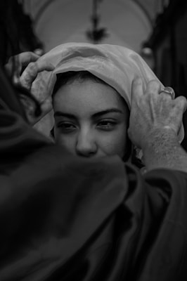 A person is having a veil or fabric draped over their head by another person in an intimate, close-up setting. The focus is on the expressive eyes of the individual receiving the veil.