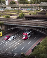 An urban multi-level infrastructure, featuring an overhead railway bridge with graffiti, a lower road with a tram and two cars traveling below, surrounded by greenery and industrial buildings in the background.