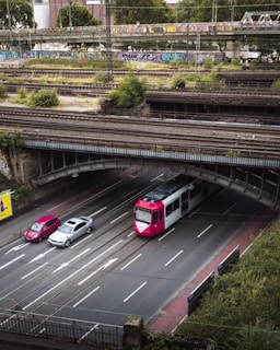 An urban multi-level infrastructure, featuring an overhead railway bridge with graffiti, a lower road with a tram and two cars traveling below, surrounded by greenery and industrial buildings in the background.
