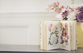An open book displaying botanical illustrations of colorful flowers on both pages. Next to the book is a yellow vase filled with a bouquet of various flowers, including pink, white, and purple blooms. The background is softly blurred, giving a serene and cozy atmosphere.