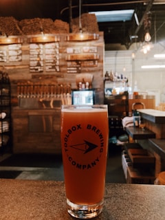 A glass of beer from Toolbox Brewing Company is placed on a counter in a rustic bar setting. Behind the glass, there is a wall with wooden accents and a row of beer taps. A menu board is visible in the background, and soft lighting gives the space a warm ambiance.