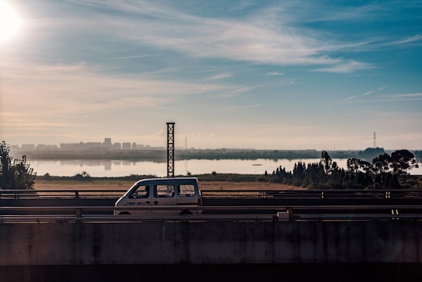 A small white van drives along an elevated highway with a scenic backdrop of a city skyline under a clear blue sky. The view includes a river and distant buildings, with trees and greenery in the foreground.