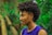 A person with curly hair styled in an afro mohawk and intricate braids on the side is shown in profile view. They are wearing a sleeveless purple top and a silver chain necklace. The background is a lush green forest, slightly blurred to emphasize the subject.