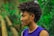 A person with curly hair styled in an afro mohawk and intricate braids on the side is shown in profile view. They are wearing a sleeveless purple top and a silver chain necklace. The background is a lush green forest, slightly blurred to emphasize the subject.