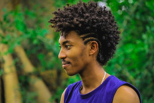 A person with curly hair styled in an afro mohawk and intricate braids on the side is shown in profile view. They are wearing a sleeveless purple top and a silver chain necklace. The background is a lush green forest, slightly blurred to emphasize the subject.