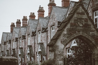 A row of traditional stone houses with steep, slate roofs and prominent brick chimneys. The architectural style features pointed gables and uniform windows. The foreground includes an arched stone structure with intricate details.