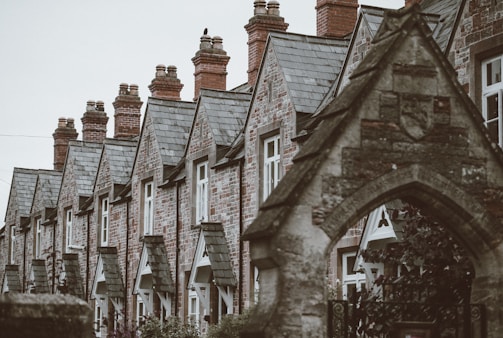A row of traditional stone houses with steep, slate roofs and prominent brick chimneys. The architectural style features pointed gables and uniform windows. The foreground includes an arched stone structure with intricate details.