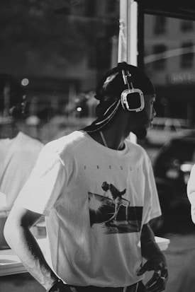 A person wearing a t-shirt with a printed graphic and headphones stands in an urban environment. The image is in black and white, adding a nostalgic or timeless feel. Reflections can be seen in the window behind the person.