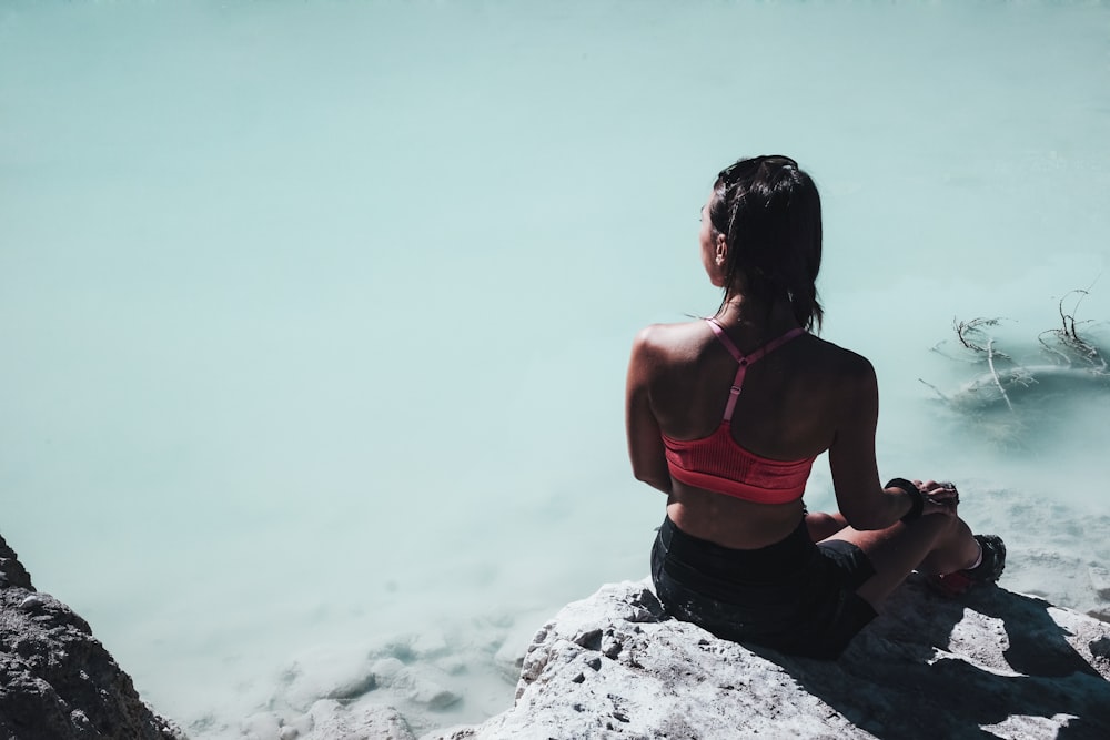 woman in pink sports bra sitting on rock near body of water at daytime