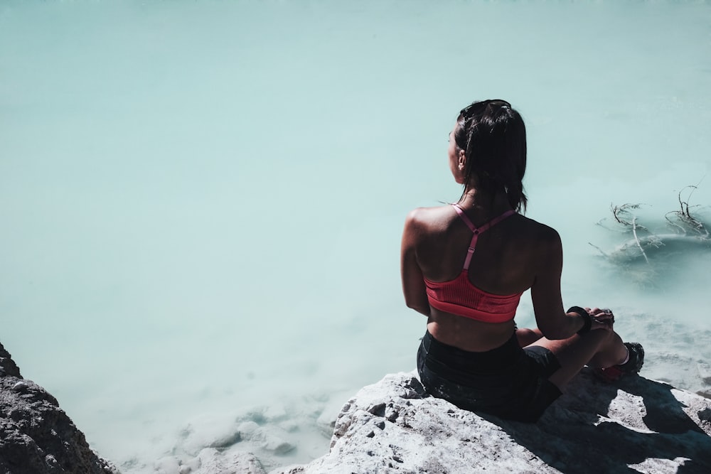 Mujer en sujetador deportivo rosa sentada en la roca cerca del cuerpo de agua durante el día