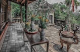 A serene patio with a traditional aesthetic features two decorative vases with yellow flowers on a wooden table. Nearby, an ornate incense burner stands on a tiled floor. In the background, lush green foliage and an ancient stone structure are visible.
