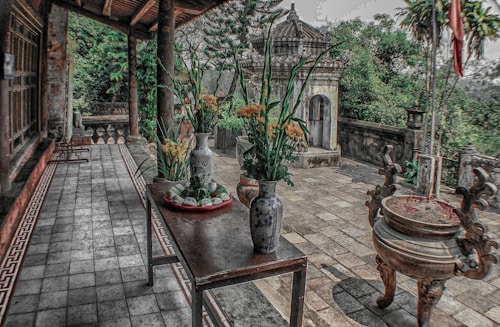 A serene patio with a traditional aesthetic features two decorative vases with yellow flowers on a wooden table. Nearby, an ornate incense burner stands on a tiled floor. In the background, lush green foliage and an ancient stone structure are visible.