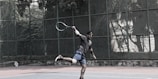 A person in sports attire is playing tennis on an outdoor court surrounded by a green metal fence and dense trees in the background. The player is mid-action, holding a tennis racket and displaying a dynamic, athletic pose. The court surface is a muted reddish color with a lighter, worn area at the foreground.
