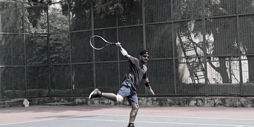 A person in sports attire is playing tennis on an outdoor court surrounded by a green metal fence and dense trees in the background. The player is mid-action, holding a tennis racket and displaying a dynamic, athletic pose. The court surface is a muted reddish color with a lighter, worn area at the foreground.