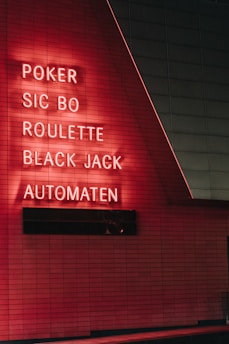 Red neon letters are mounted on a dark tiled wall, displaying the words Poker, Sic Bo, Roulette, Black Jack, and Automaten.