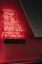 Red neon letters are mounted on a dark tiled wall, displaying the words Poker, Sic Bo, Roulette, Black Jack, and Automaten.