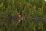 A small red cabin nestled among dense green trees is reflected in a calm lake. The scene is peaceful with lush foliage and clear water, highlighting the solitude and tranquility of nature.