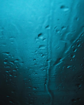 Water droplets and streaks move down a smooth surface, possibly glass, with a blue-green tint. The light gives the surface a shimmering appearance, creating a sense of wetness and fluidity.