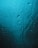 Water droplets and streaks move down a smooth surface, possibly glass, with a blue-green tint. The light gives the surface a shimmering appearance, creating a sense of wetness and fluidity.
