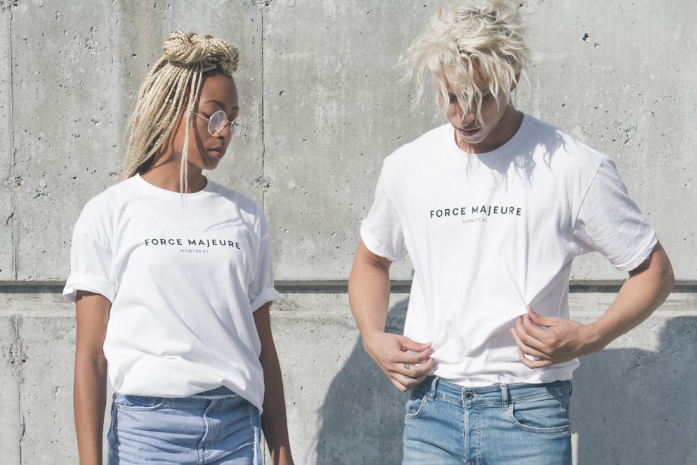 Homme et femme en t-shirts à col rond blancs et gris imprimés Force Majeure debout près d’un mur de béton gris