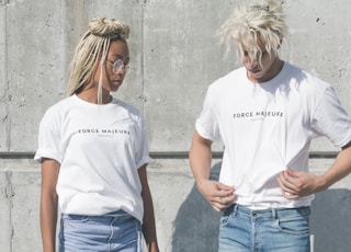 man and woman in white-and-gray force majeure-printed crew-neck t-shirts standing near gray concrete wall