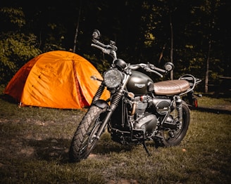 A motorcycle is parked on grass with a bright orange tent behind it. The background consists of dense, dark trees casting shadows. The motorcycle has a classic design with black and metal elements.