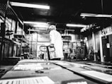 A person wearing a white lab coat and hairnet stands in an industrial setting, likely a warehouse or processing facility. The individual is holding onto a hand truck or dolly, surrounded by large boxes and shelving units. The lighting is stark and the black-and-white filter adds a vintage or gritty aesthetic to the scene, emphasizing the utilitarian nature of the environment.