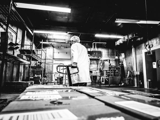 A person wearing a white lab coat and hairnet stands in an industrial setting, likely a warehouse or processing facility. The individual is holding onto a hand truck or dolly, surrounded by large boxes and shelving units. The lighting is stark and the black-and-white filter adds a vintage or gritty aesthetic to the scene, emphasizing the utilitarian nature of the environment.