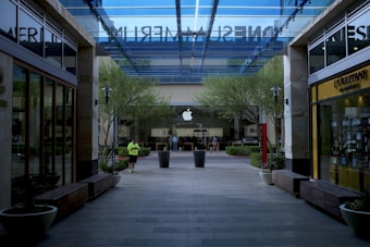 A modern shopping center with a glass canopy overhead, displaying the entrance to an Apple store at the center. Flanked by two L'OCCITANE outlets on the sides, with trees and planters adding greenery. A person in bright athletic clothing stands in the middle walkway.