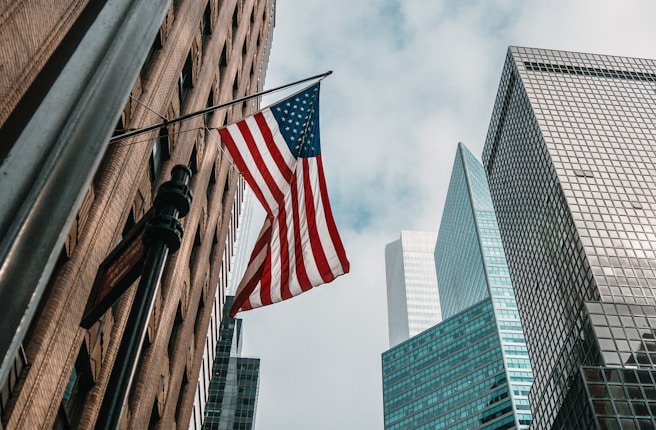 An American flag flutters against a backdrop of towering skyscrapers under a partly cloudy sky. The perspective from below accentuates the height of the buildings, giving a sense of urban grandeur.