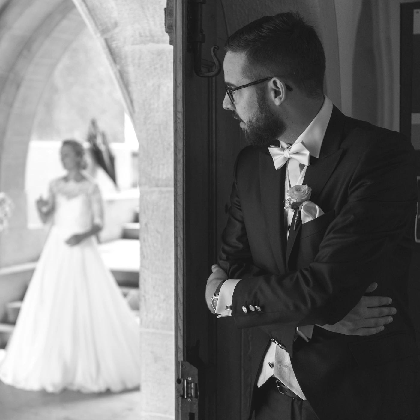 Black and white wedding photograph of the groom with his back turned while the bride approaches from behind in a first look moment
