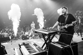 A musician energetically performs on stage with a guitar and electronic keyboards while an enthusiastic crowd watches. Smoke effects are visible, adding to the dynamic atmosphere. The stage setup includes various electronic instruments and a laptop.