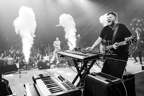 A musician energetically performs on stage with a guitar and electronic keyboards while an enthusiastic crowd watches. Smoke effects are visible, adding to the dynamic atmosphere. The stage setup includes various electronic instruments and a laptop.