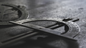 A close-up shot of two metal protractors with engraved measurement markings. They are laid on a textured surface with soft lighting creating shadows, enhancing the industrial and mechanical look.