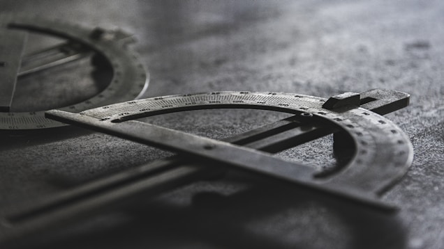 A close-up shot of two metal protractors with engraved measurement markings. They are laid on a textured surface with soft lighting creating shadows, enhancing the industrial and mechanical look.